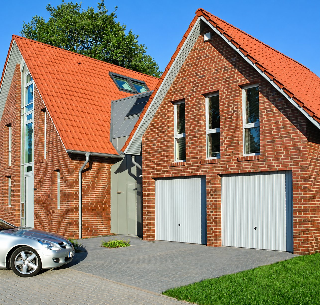 Maison moderne en briques rouges à toit de tuiles orange, équipée de deux portes de garage blanches, avec une voiture grise garée sur l'allée pavée.
