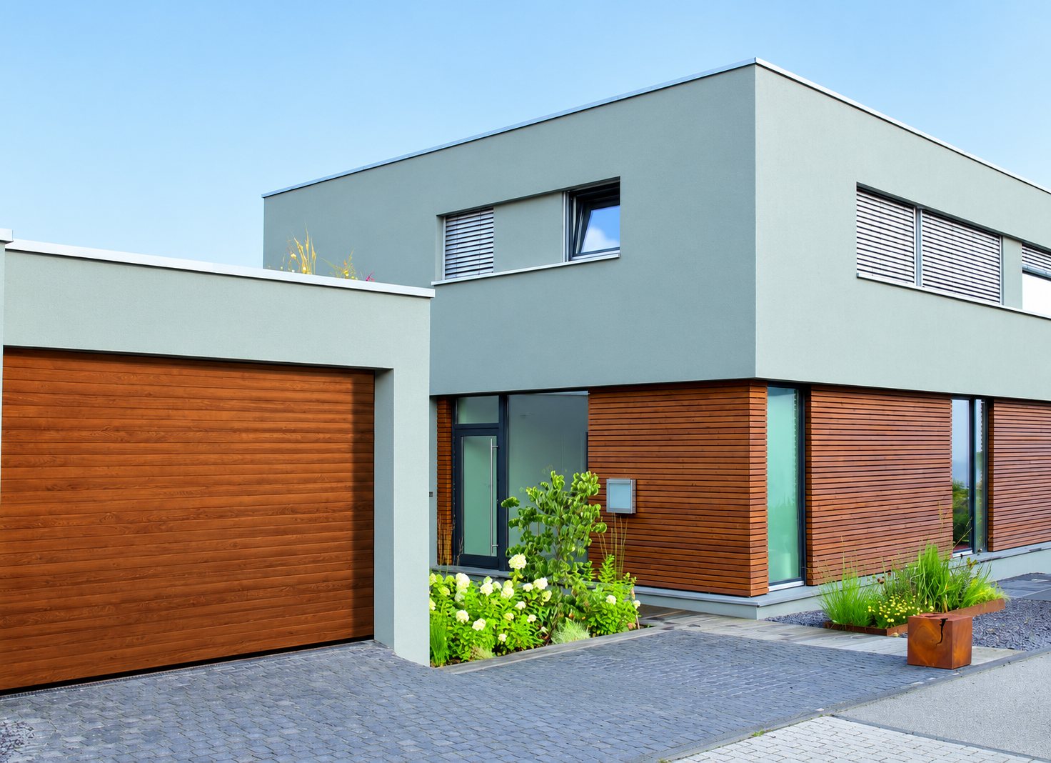 Façade d'une maison contemporaine à toit plat avec une grande porte de garage couleur bois, des bardages en bois et un petit jardin fleuri devant l'entrée.