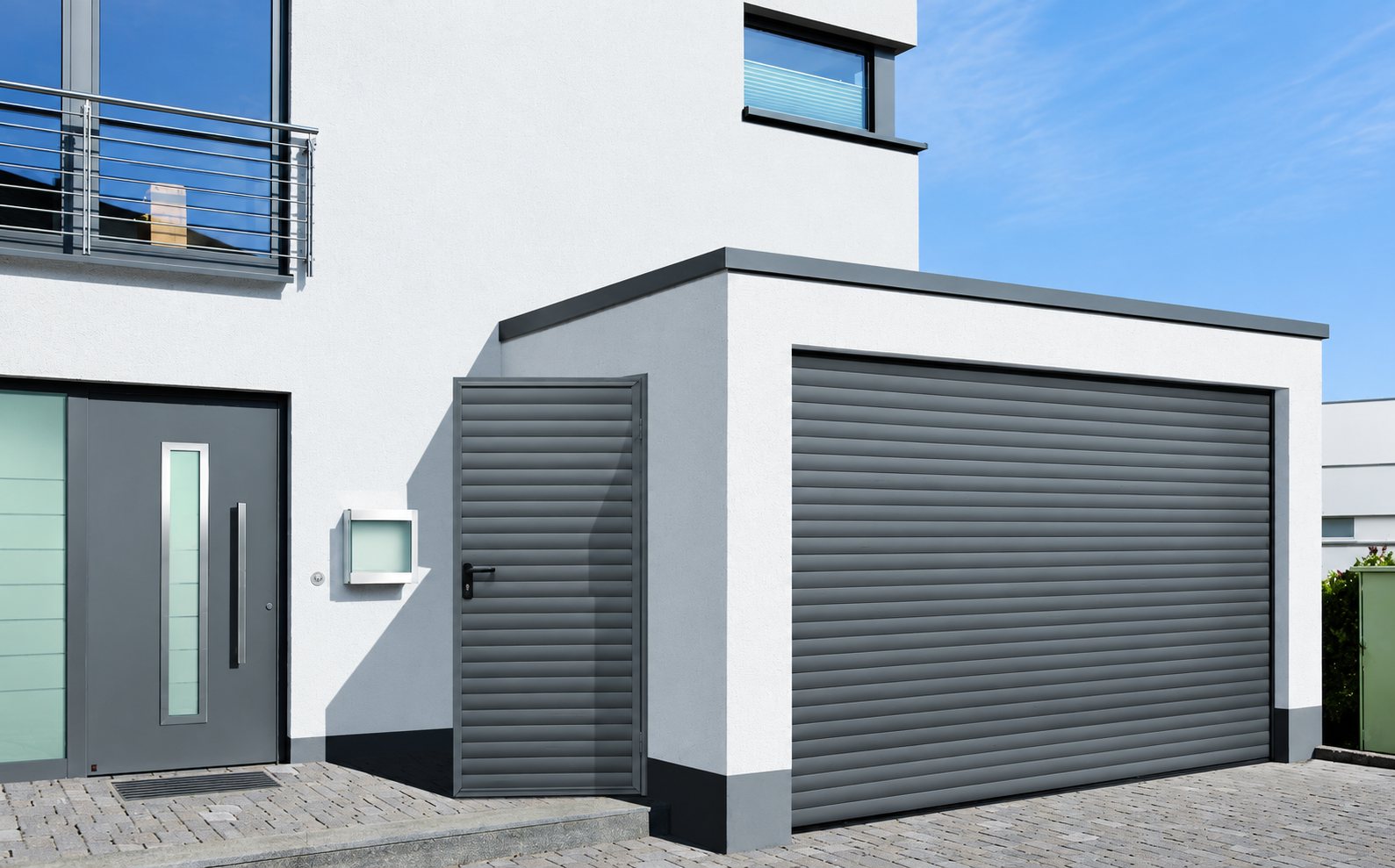 Façade blanche d'une maison moderne avec une porte de garage enroulable grise, une porte latérale assortie et une porte d'entrée en aluminium.