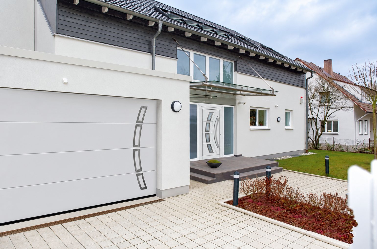 Façade d'une maison moderne blanche avec une porte de garage sectionnelle blanche à motifs en inox et une porte d'entrée assortie, devant une allée pavée.