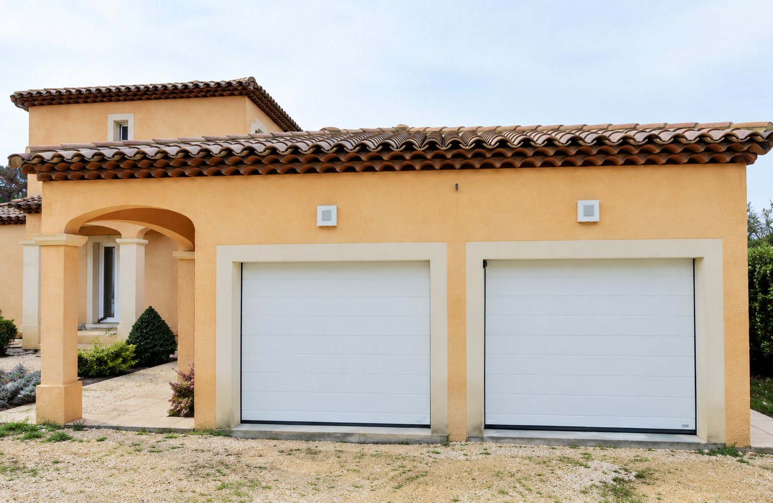 Façade d'une maison à l'enduit orange avec un toit de tuiles et deux portes de garage blanches côte à côte.