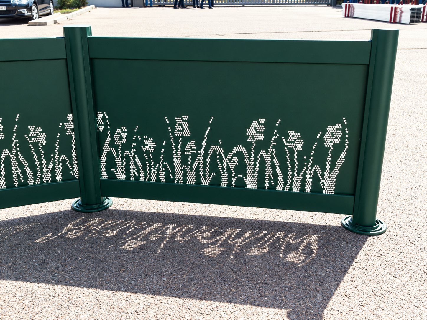 Un panneau de clôture métallique vert, perforé de motifs de fleurs et d'herbes, projette son ombre ajourée sur le sol d'un parking ensoleillé.