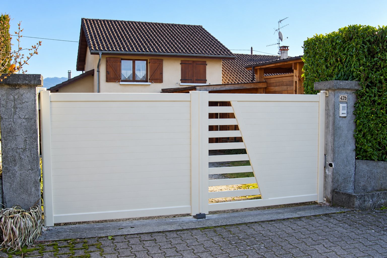 Un portail battant beige à lames horizontales avec une partie ajourée en diagonale, installé entre deux piliers en béton devant une maison.