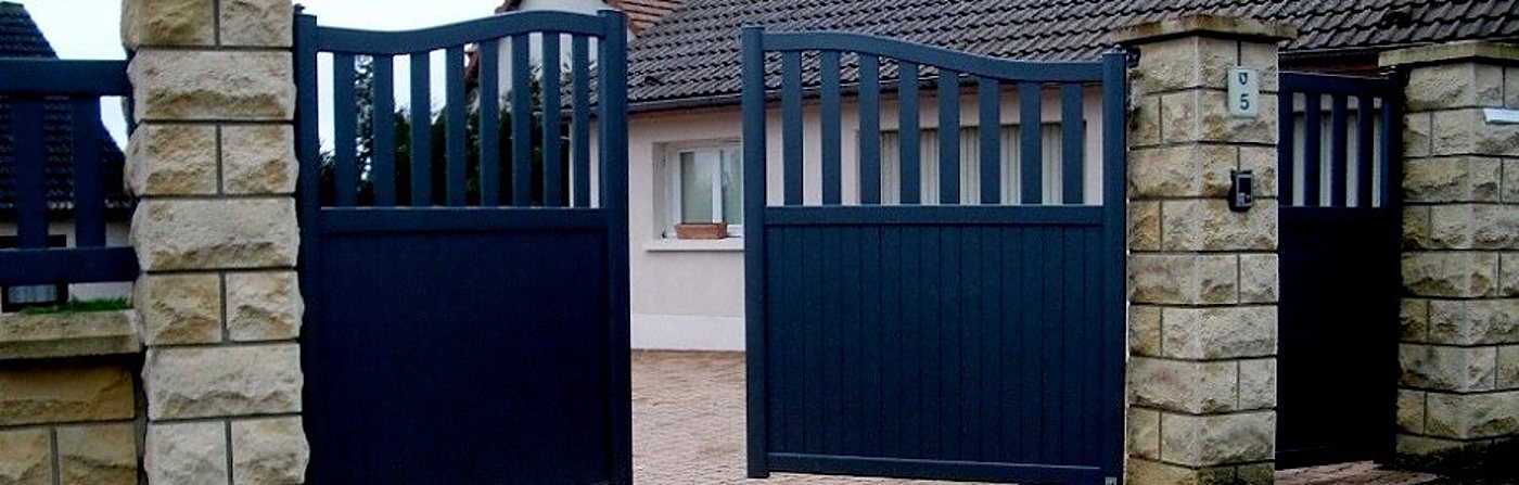 Un portail battant en aluminium bleu anthracite à deux vantaux, ouvert entre des piliers en pierre claire, devant une maison au toit de tuiles.