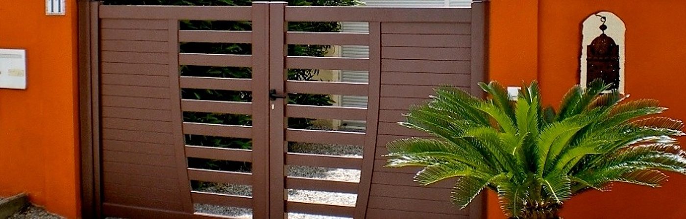 Un portail battant brun à lames horizontales ajourées est installé entre deux murs orange, avec un palmier cycas au premier plan.