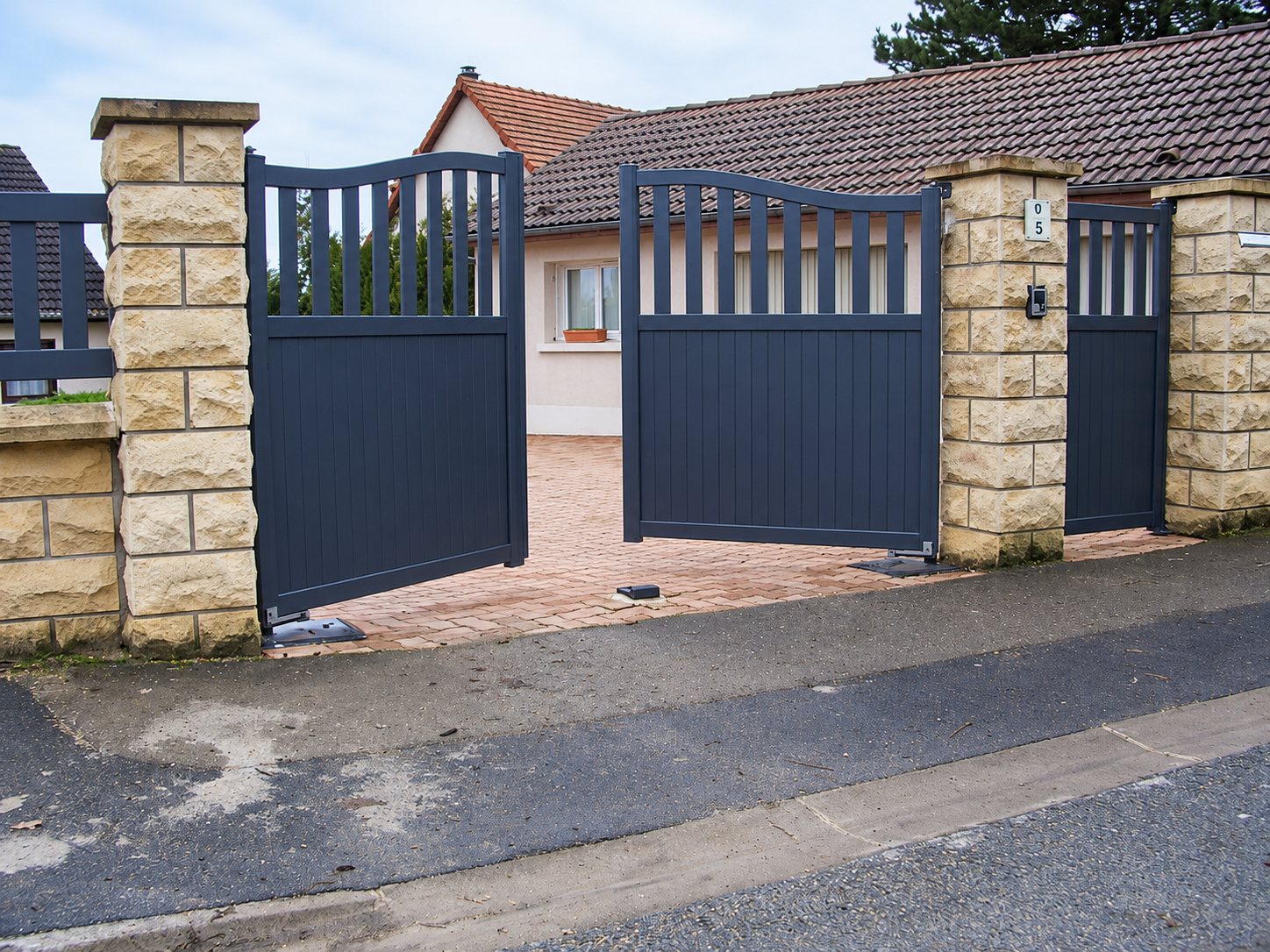 Un portail battant en aluminium gris anthracite, entrouvert, encadré de piliers en pierre claire à l'entrée d'une maison avec allée pavée.
