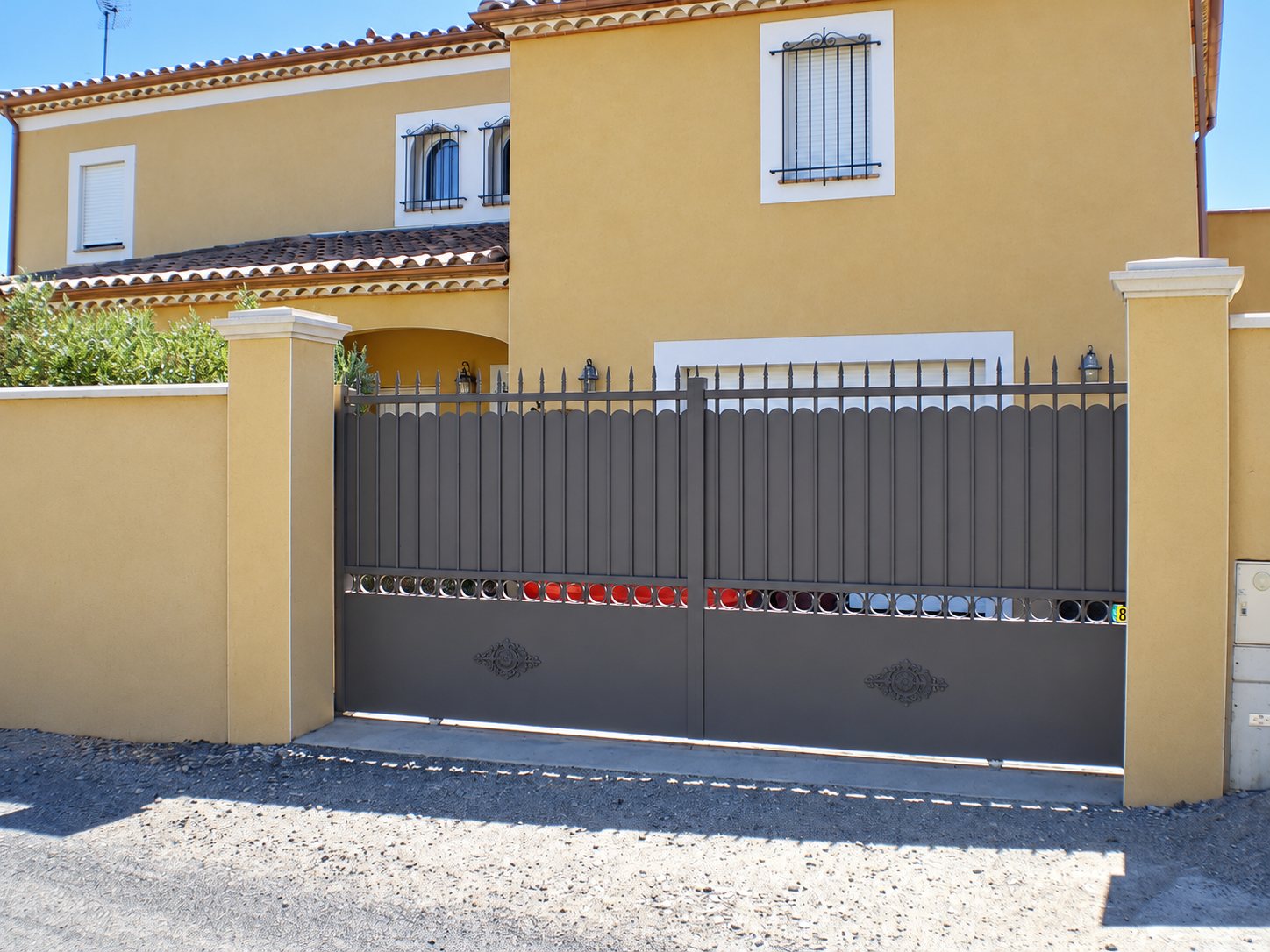 Un grand portail battant marron à barreaux et motifs décoratifs, encadré par deux piliers ocre devant une maison méditerranéenne.