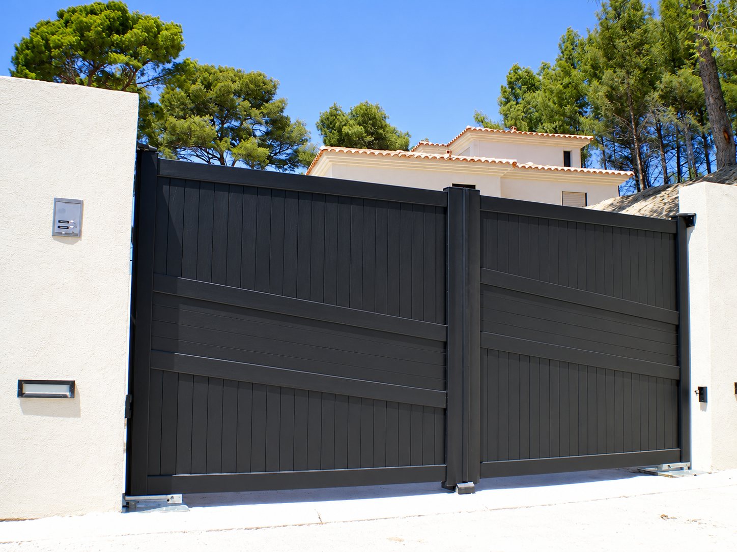 Un grand portail battant noir à lames horizontales et verticales entre deux murs blancs, avec une villa à toit de tuiles et des pins en arrière-plan.