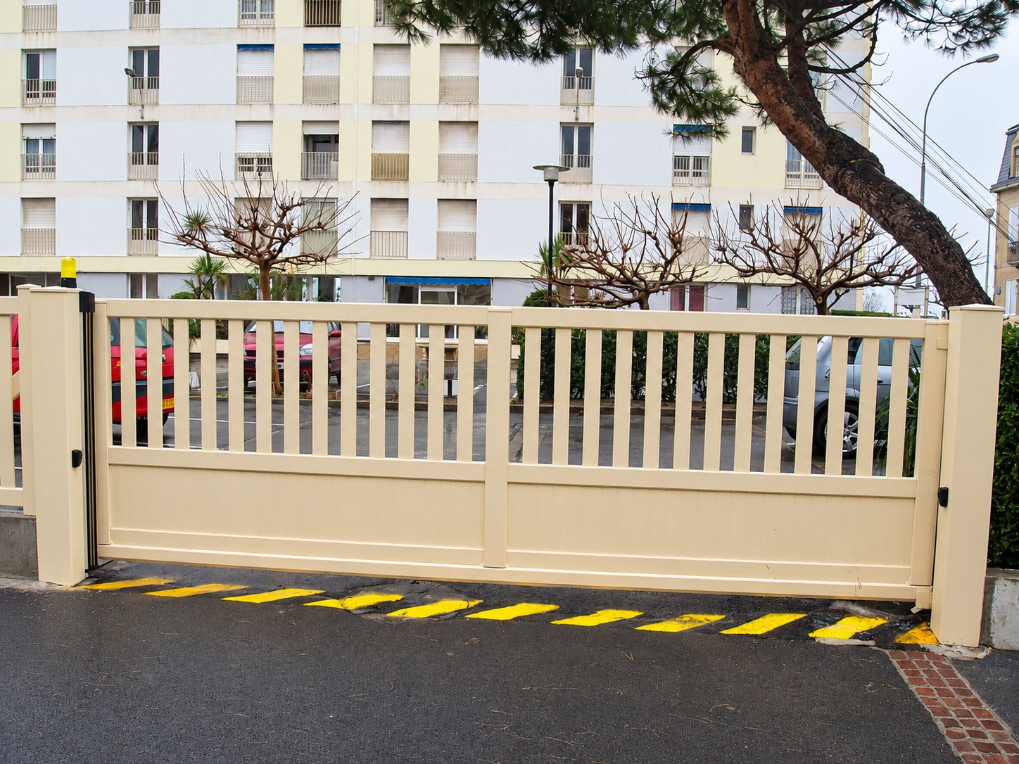 Un grand portail beige à barreaux verticaux ferme l'entrée d'une résidence, avec des bandes jaunes peintes au sol et un immeuble en arrière-plan.