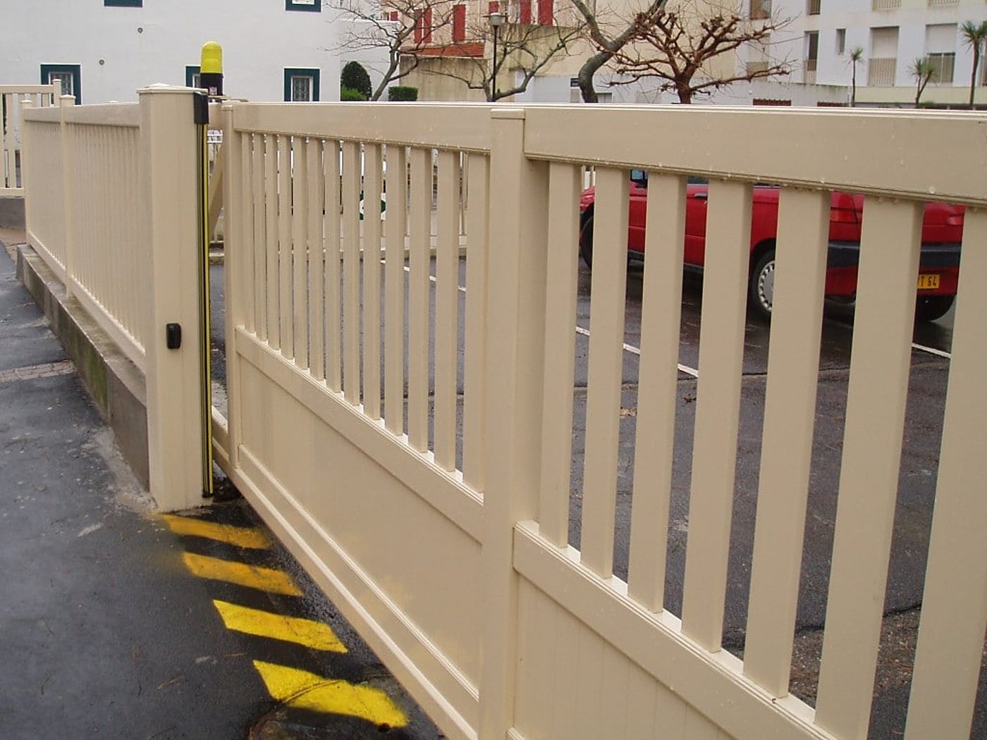 Portail coulissant beige à barreaux verticaux avec feu clignotant jaune et marquage jaune et noir au sol, devant un parking où stationne une voiture rouge.