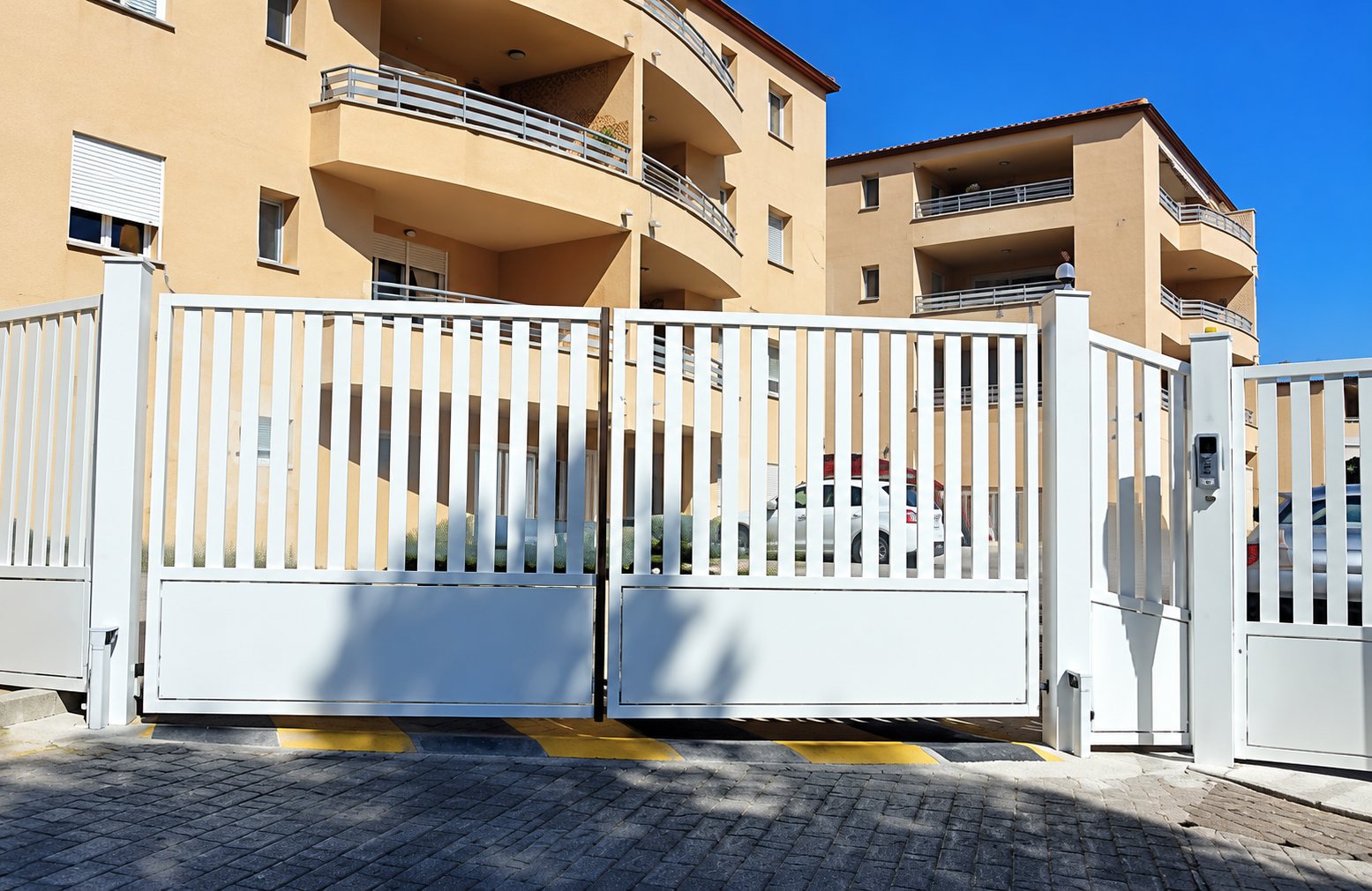 Un grand portail blanc à lames verticales ferme l'entrée d'une résidence aux façades ocre avec balcons.
