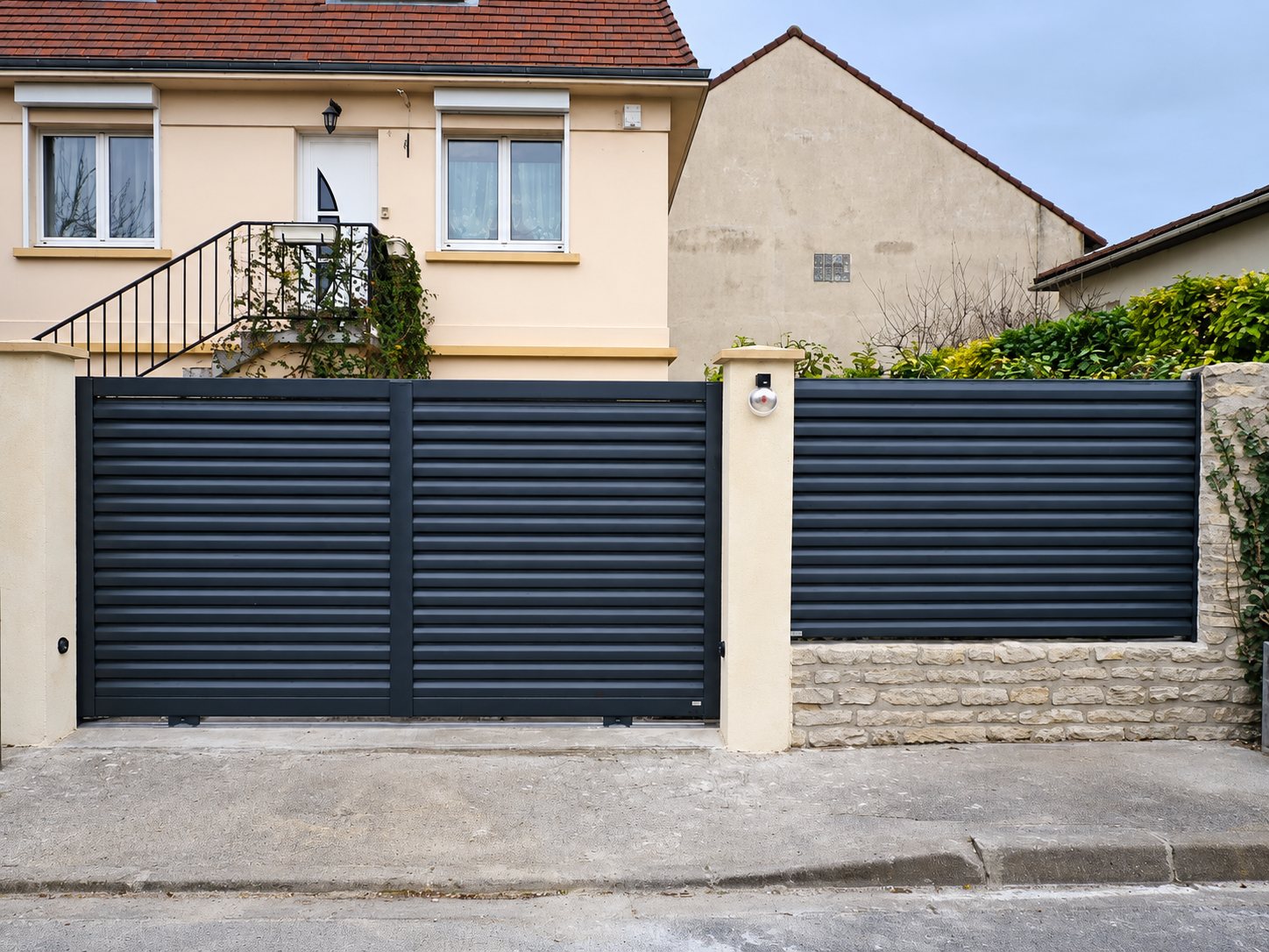 Portail coulissant et clôture à lames horizontales gris anthracite installés entre des piliers en pierre claire devant une maison rose.