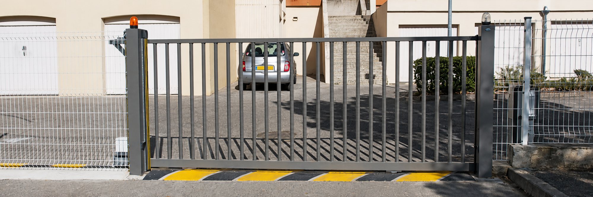 Un portail coulissant gris à barreaux verticaux, équipé d'un feu orange, ferme l'accès d'un parking résidentiel où une petite voiture grise est garée.