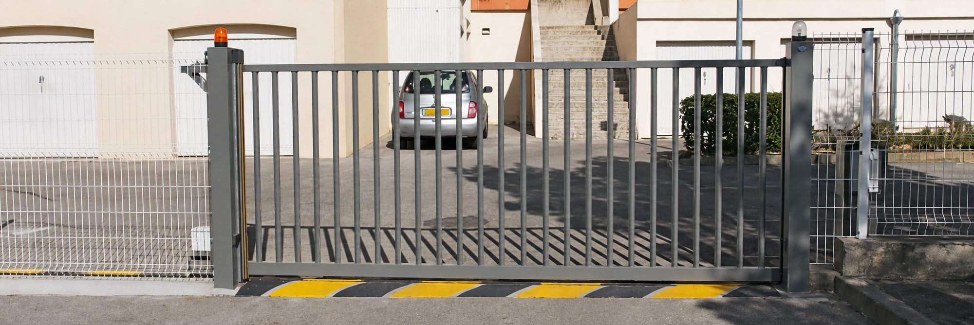 Un portail coulissant métallique gris à barreaux verticaux ferme l'entrée d'une résidence, avec une voiture garée derrière et des garages en arrière-plan.