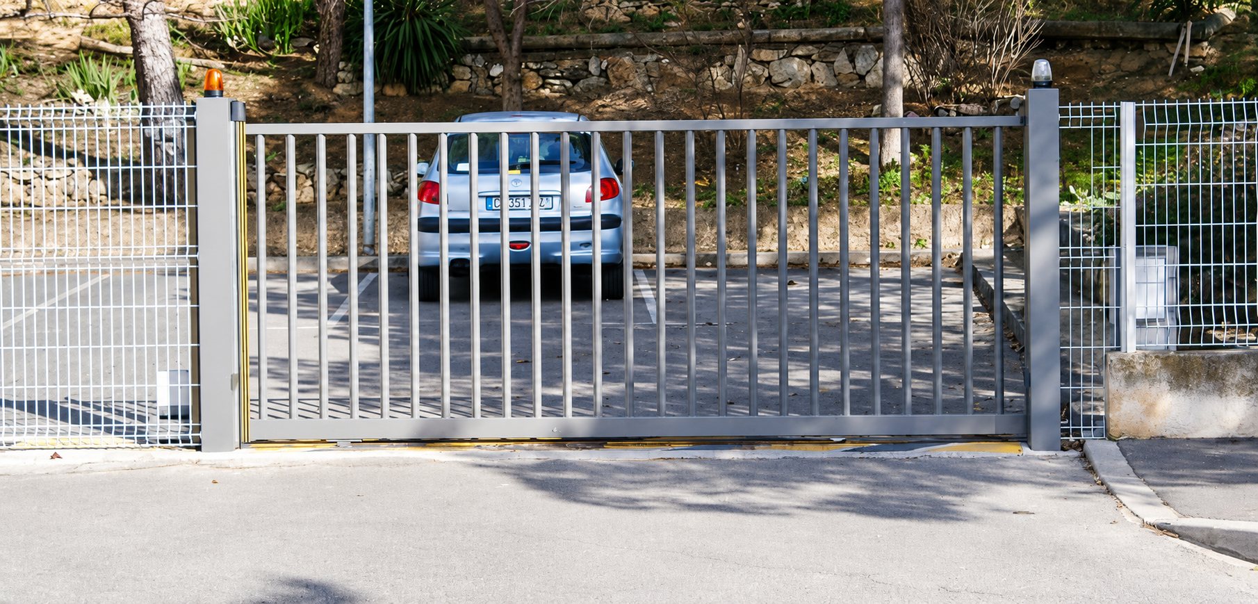 Un portail coulissant gris à barreaux verticaux, flanqué de clôtures grillagées blanches, laisse apercevoir une voiture blanche garée derrière.