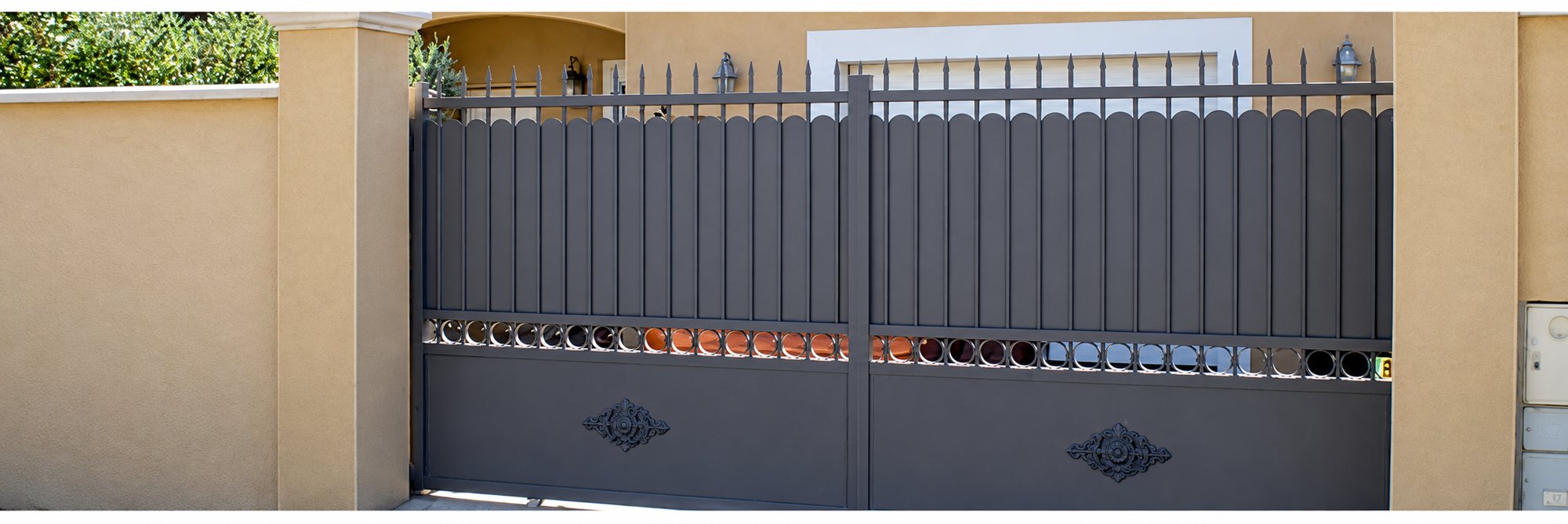 Un grand portail métallique gris à barreaux pointus et motifs décoratifs, installé entre deux piliers et des murs beiges devant une maison.