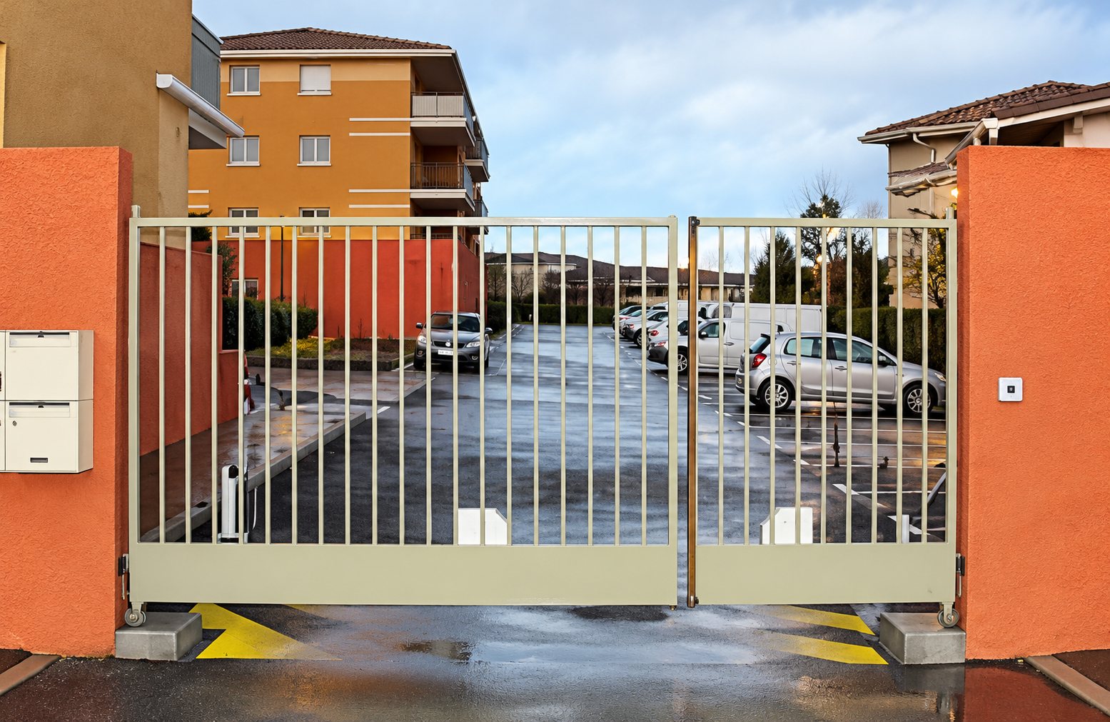 Un portail métallique à barreaux entre deux piliers orange donne sur un parking mouillé avec des voitures et un immeuble en arrière-plan.