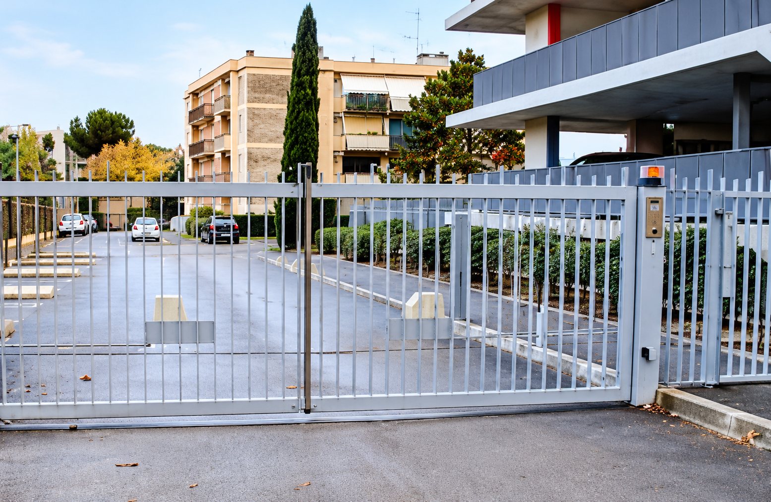 Un grand portail coulissant métallique fermé donne sur un parking bordé d'arbres et d'immeubles résidentiels.