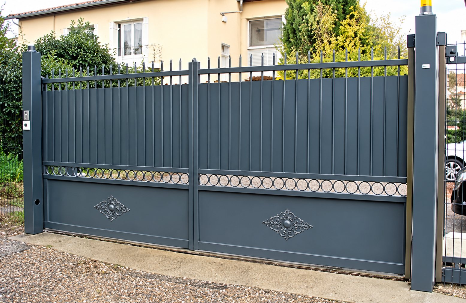 Un portail coulissant en métal gris anthracite à barreaux verticaux et motifs décoratifs, installé devant une maison et de la végétation.