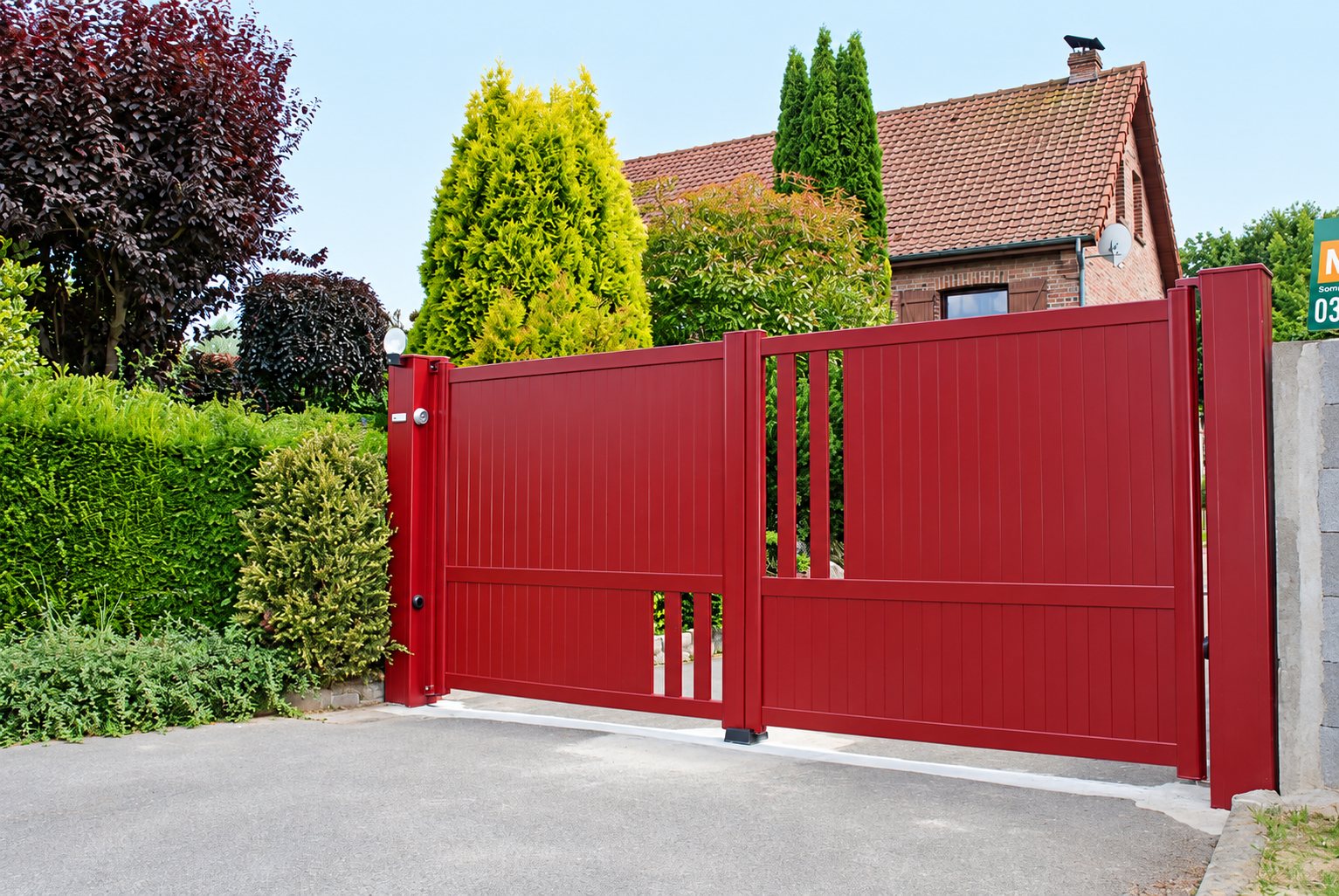 Un portail en aluminium rouge bordeaux à deux vantaux avec quelques ouvertures verticales ferme l'entrée d'une maison en briques entourée de haies et d'arbres.