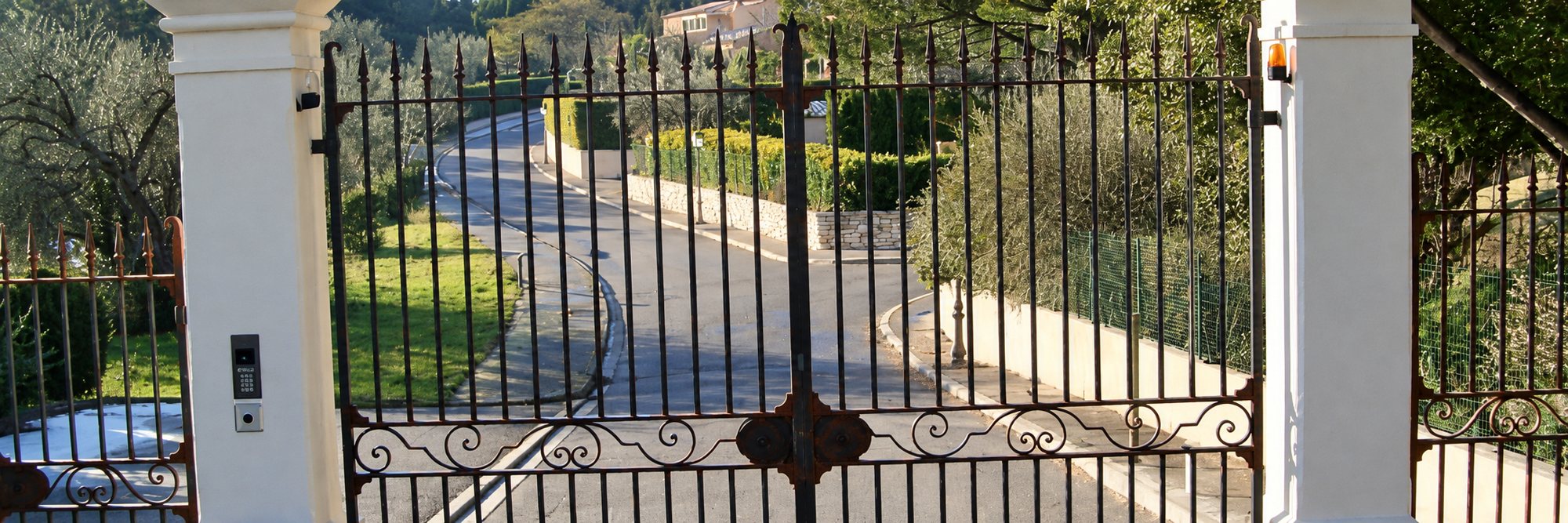 Un portail en fer forgé aux volutes décoratives, fermé entre deux piliers blancs, ouvre sur une allée qui descend vers un jardin.