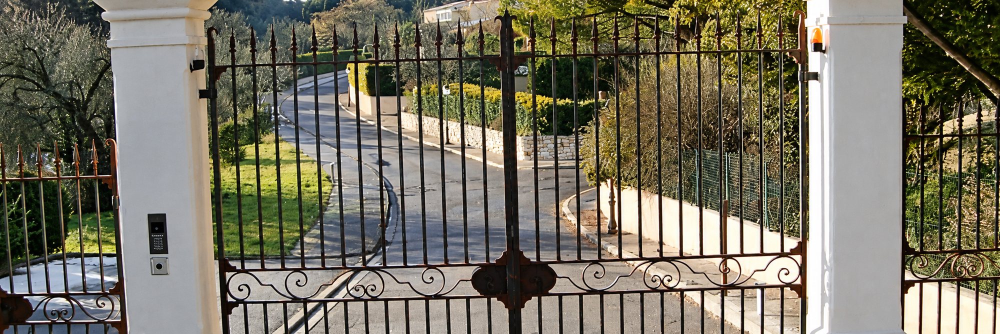 Un portail en fer forgé aux pointes de lance, encadré par deux piliers blancs, ouvre sur une route bordée de pelouse et de haies.