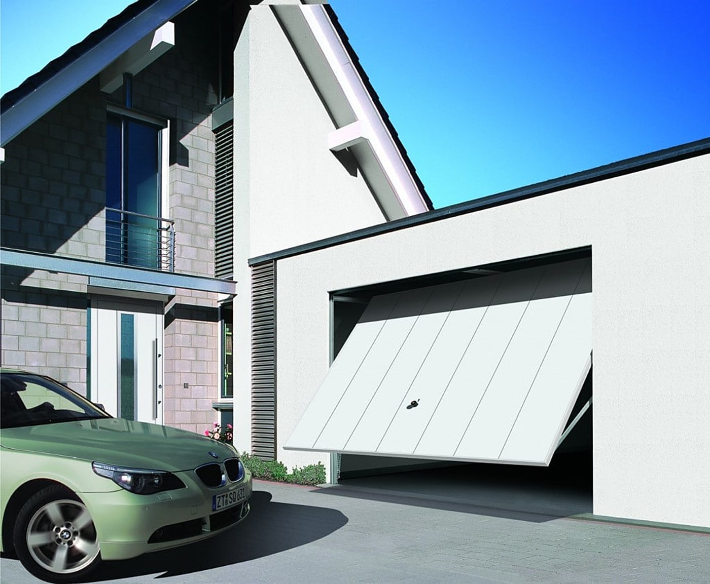 Une porte de garage basculante blanche à moitié ouverte sur une maison moderne, avec une berline verte garée devant dans l'allée.