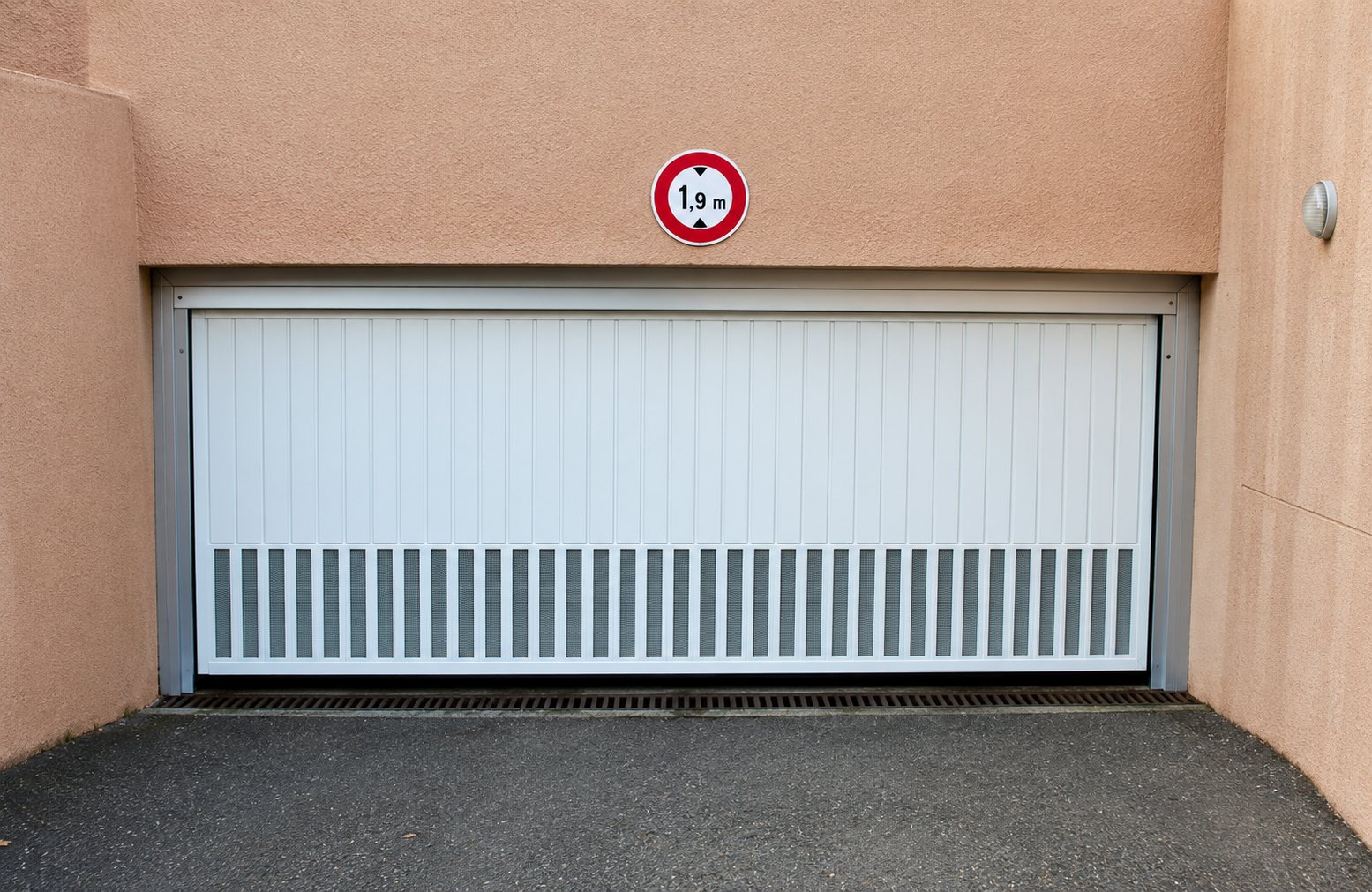 Une porte de garage blanche à lames verticales encadrée de murs beiges, surmontée d'un panneau rond de limitation.