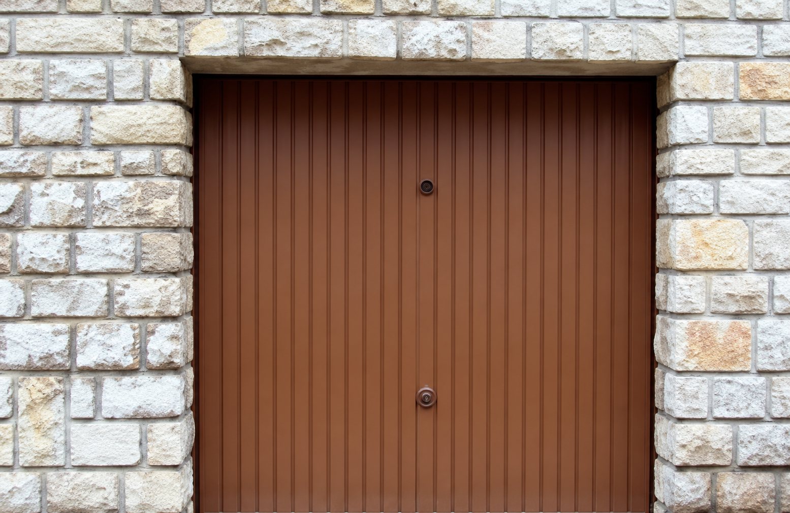 Une porte de garage sectionnelle marron à lames horizontales est encadrée par un mur en pierres de taille beiges.