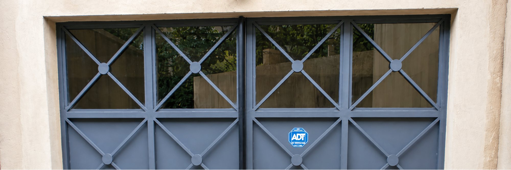 Porte de garage double en métal gris à motifs en croix, avec partie haute vitrée et un petit autocollant ADT bleu sur le battant de droite.