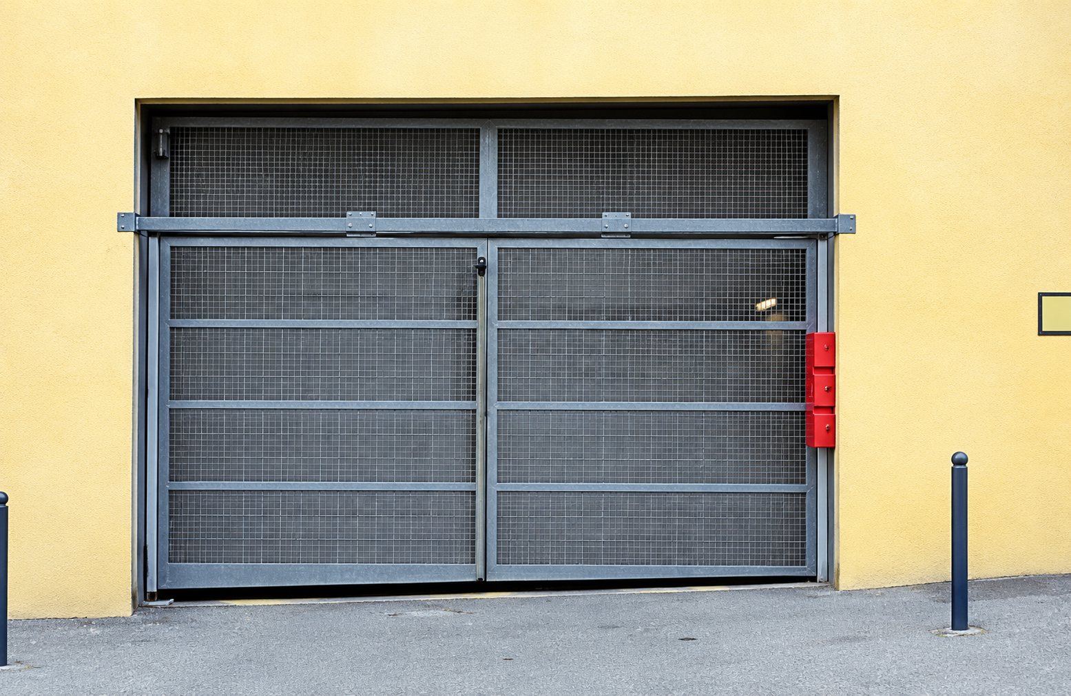Une grande porte de garage métallique grise à lames ajourées encadre l'entrée d'un bâtiment à la façade jaune, avec un extincteur rouge sur le côté.