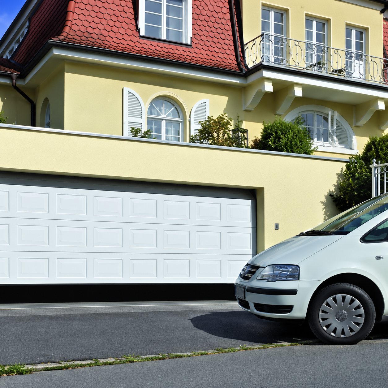Une porte de garage sectionnelle blanche installée sous une maison jaune au toit rouge, avec un monospace blanc garé devant.