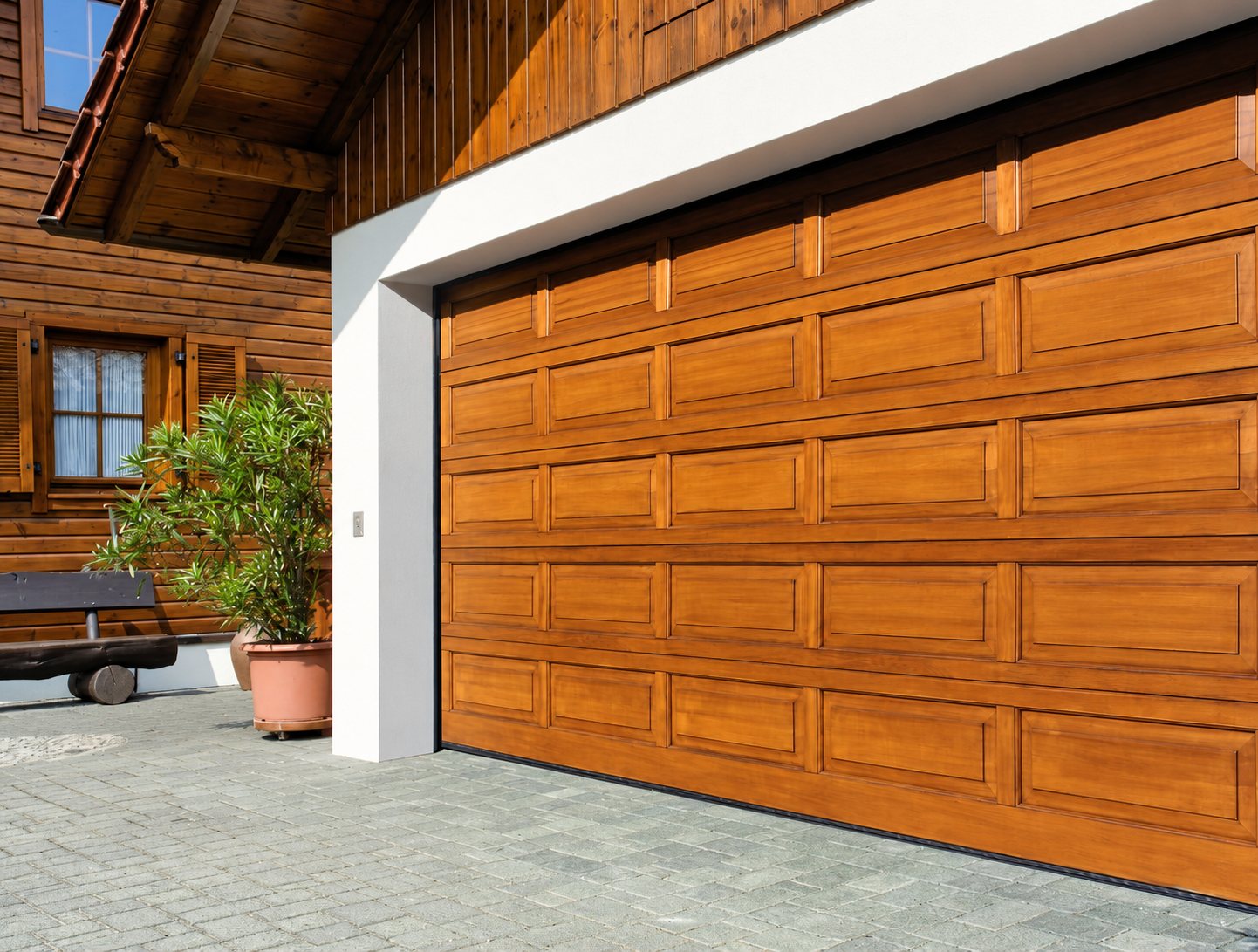 Une grande porte de garage sectionnelle en bois à panneaux, installée dans une façade blanche, devant une maison en bois avec un laurier-rose en pot.