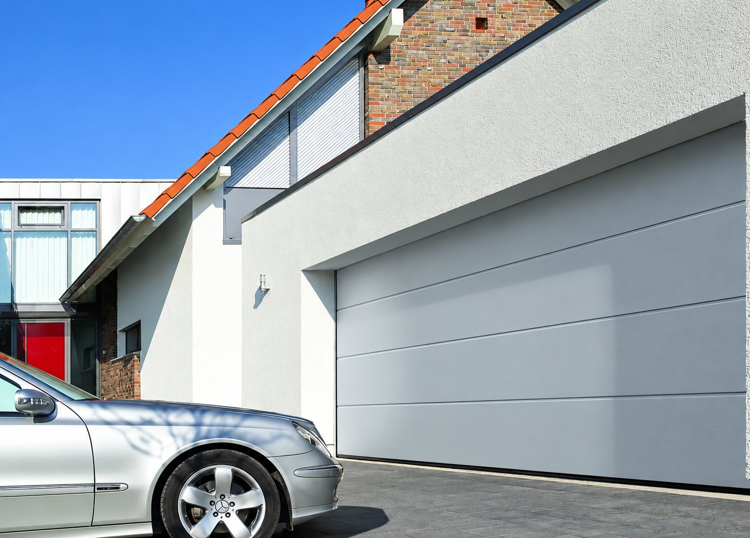 Une grande porte de garage sectionnelle grise installée sur une maison moderne en crépi blanc, avec une voiture argentée garée devant.