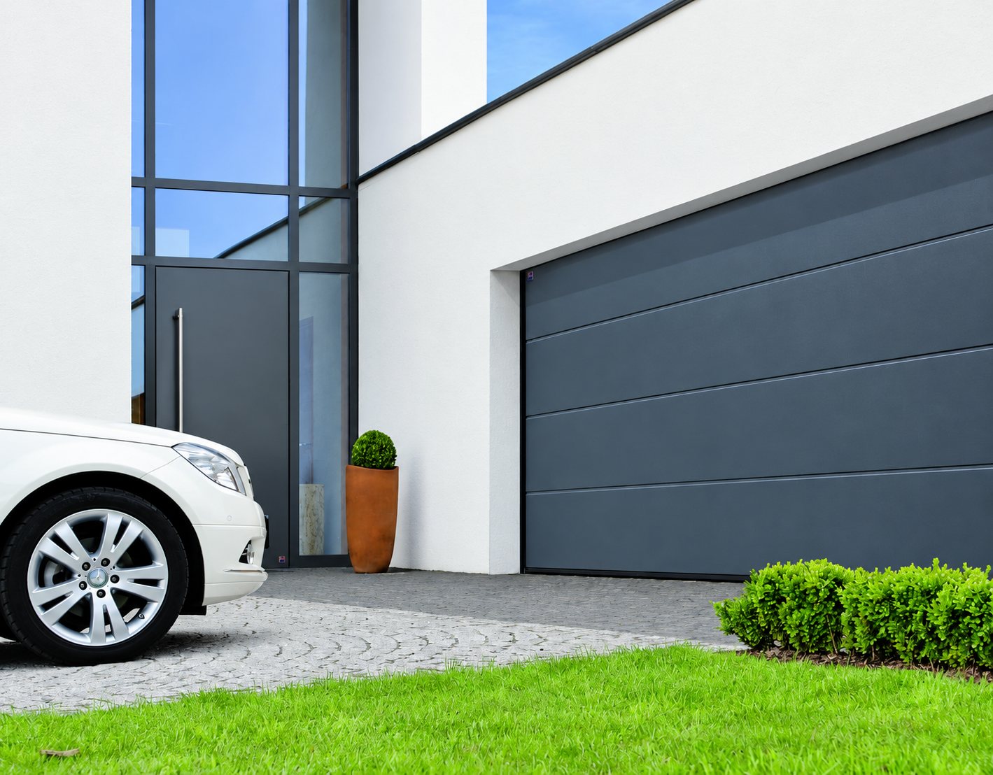 Une porte de garage sectionnelle gris anthracite sur une façade blanche moderne, avec une voiture blanche garée sur un pavage et une pelouse au premier plan.