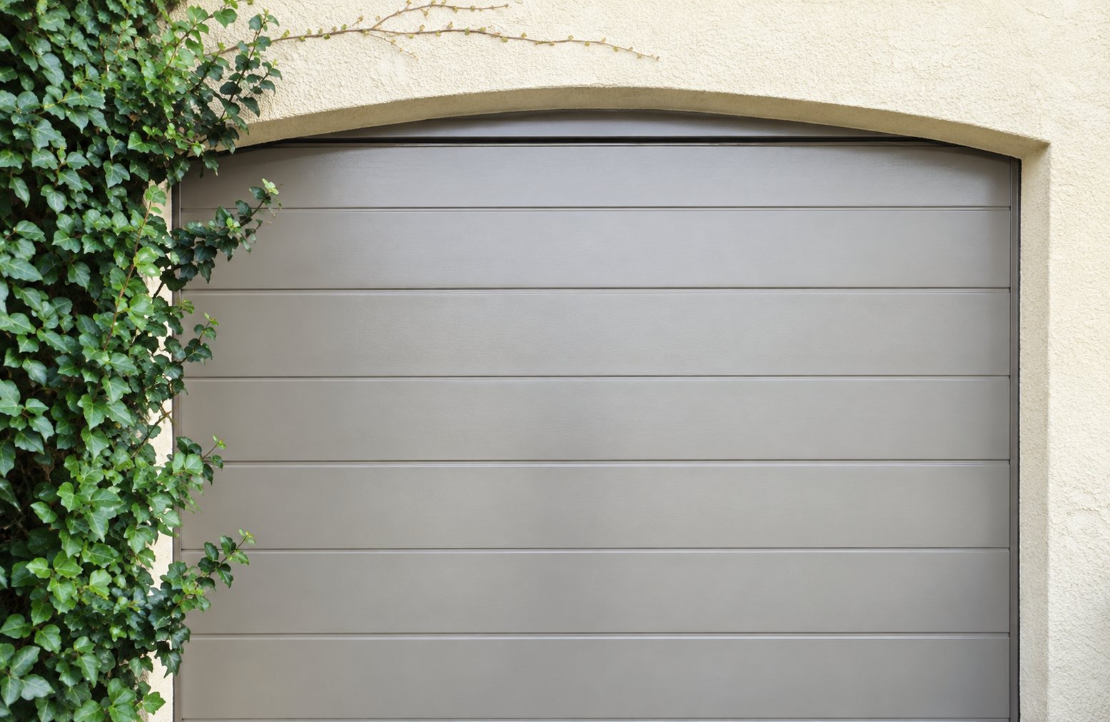 Une porte de garage sectionnelle grise à linteau arrondi, encadrée par un mur beige et du lierre sur la gauche.
