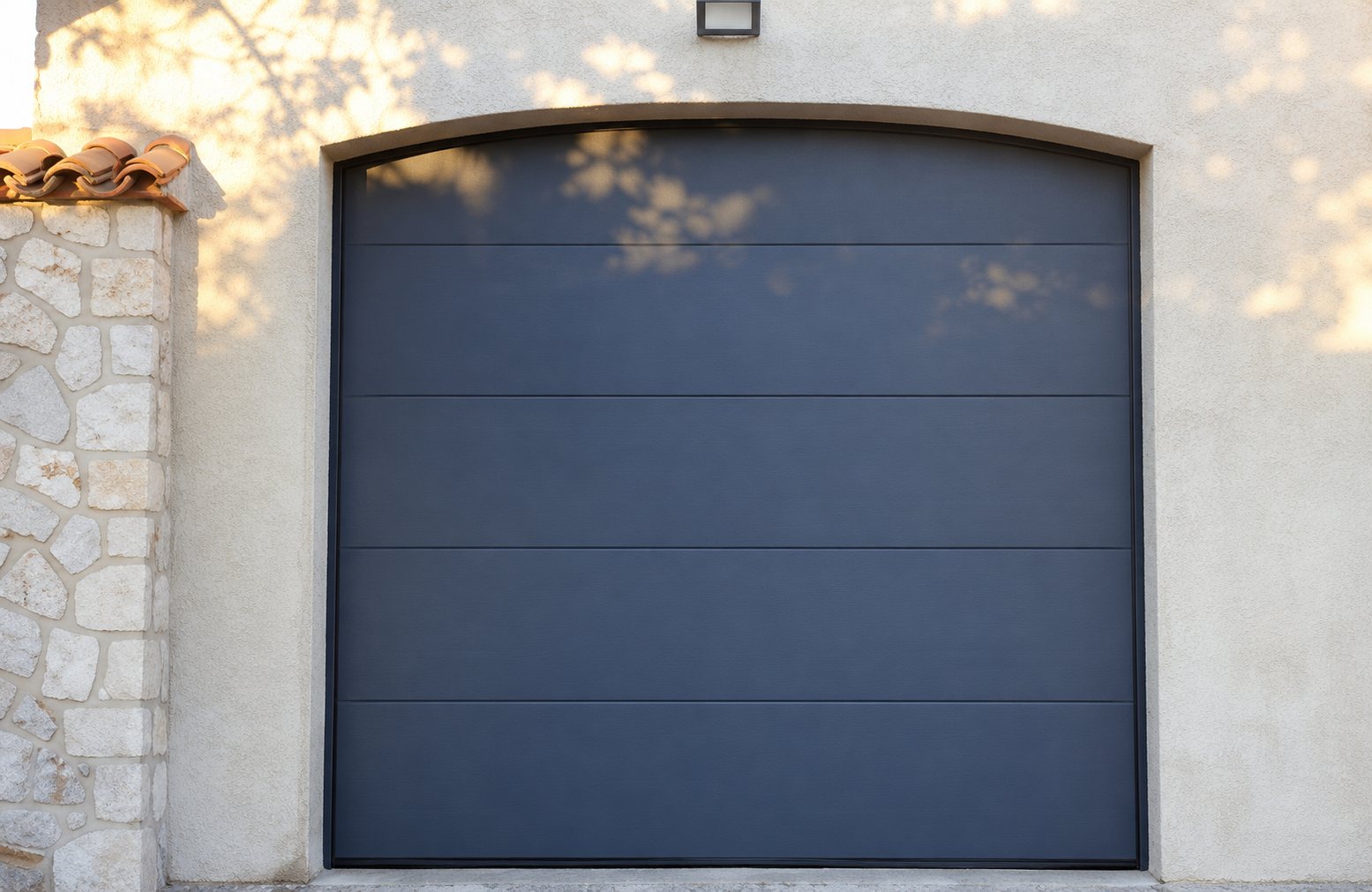 Une porte de garage sectionnelle gris anthracite à linteau cintré, posée dans une façade en crépi beige à côté d'un pilier en pierre.