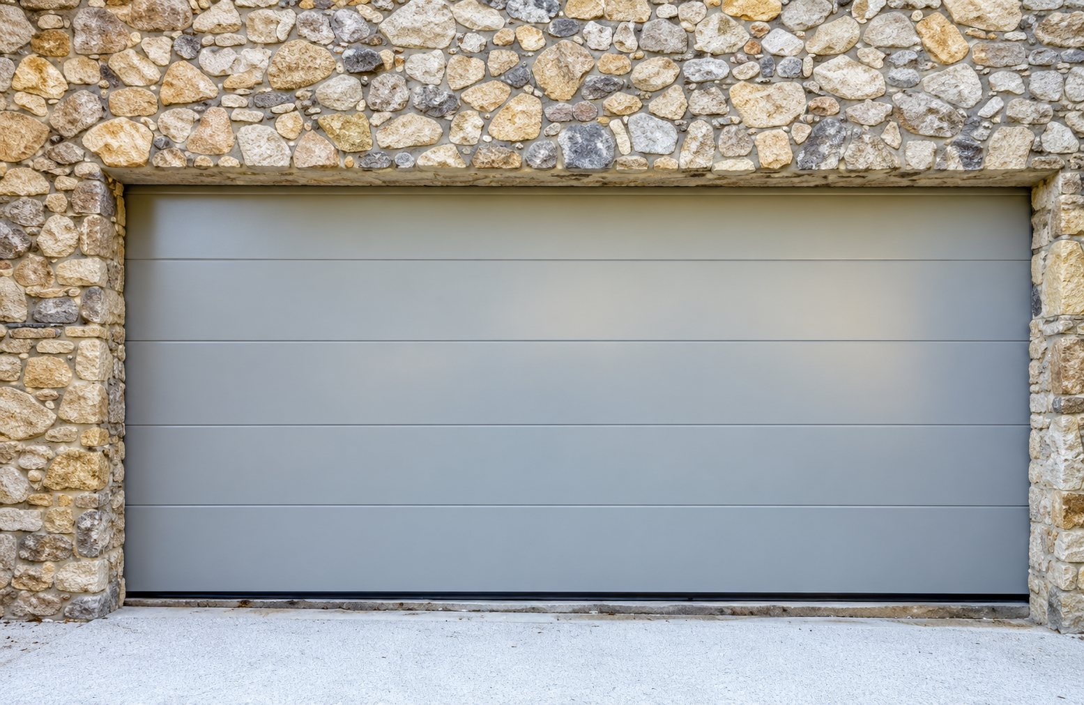 Une porte de garage sectionnelle grise est encastrée dans un mur en pierres naturelles, avec un sol en béton clair devant.