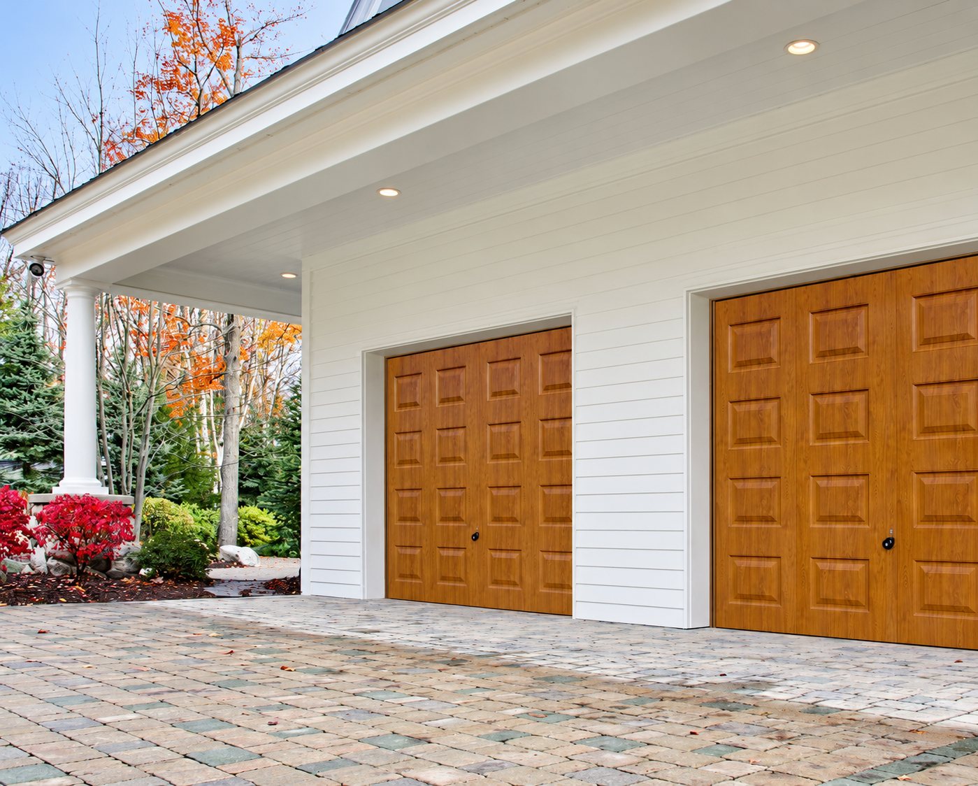 Deux portes de garage aspect bois clair à panneaux en relief installées sur une maison à façade blanche, avec une allée en pavés et un jardin d'automne.